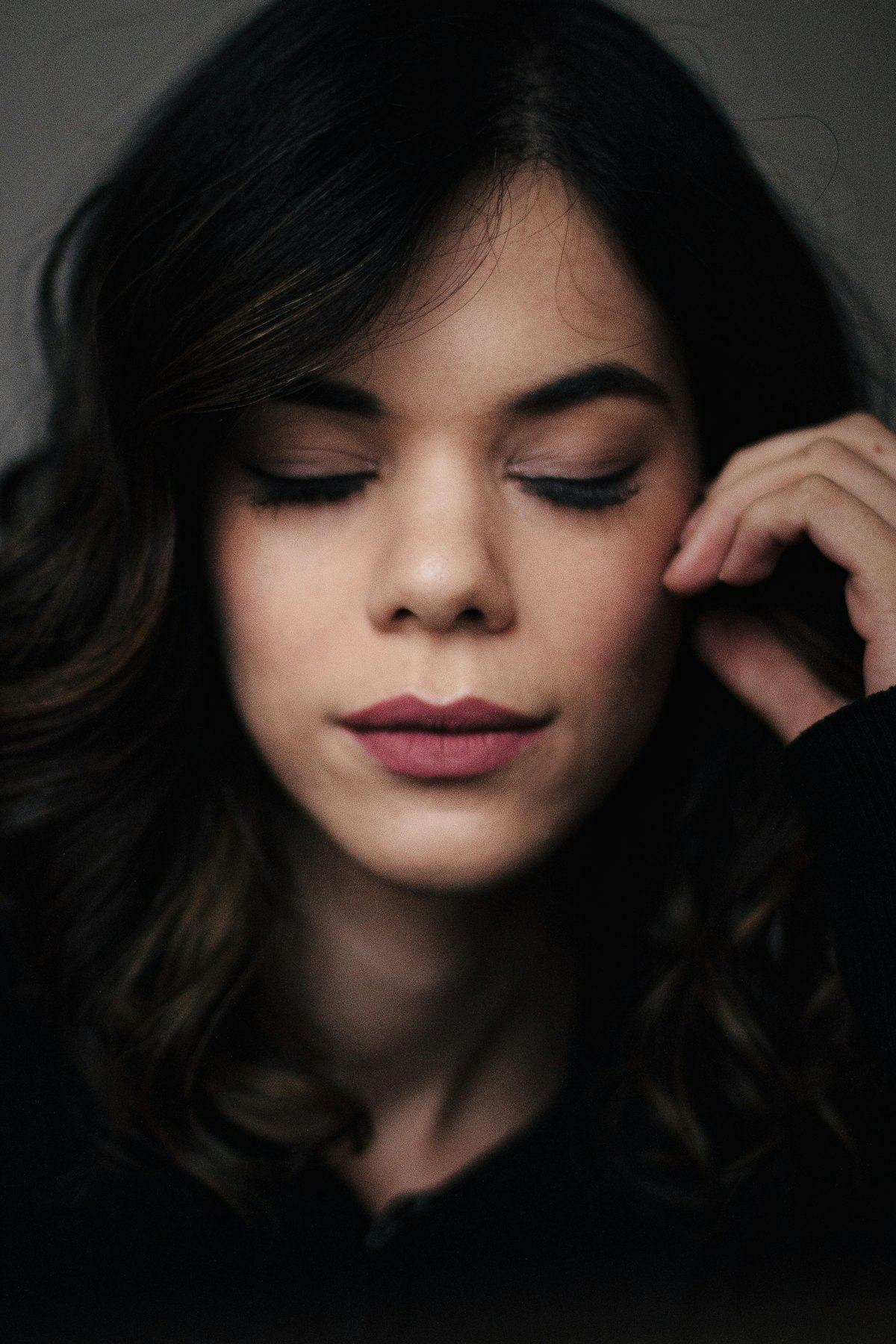 Mulher de olhos fechados, cílios longos e cabelo escuro, com a mão perto do rosto